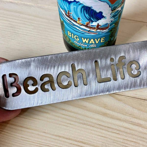 close up of metal paddle style bottle opener that says Beach Life, leaning against a bottle